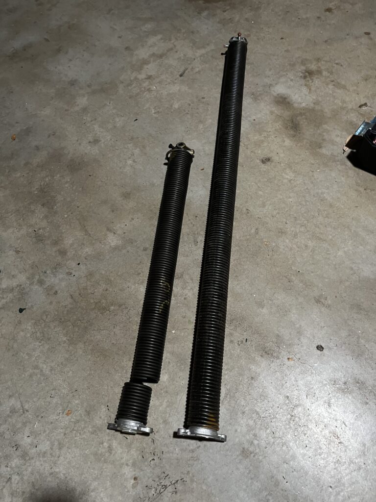 one broken garage door torsion spring laying on the floor next to a brand new one
