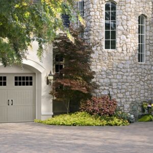 closeup of a large stone home with many trees in front and a smaller grey faux wood grain style garage door with windows on the top row