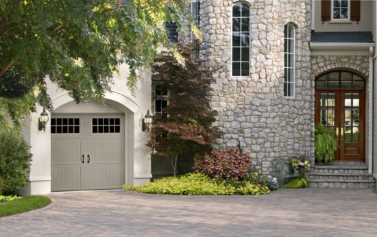closeup of a large stone home with many trees in front and a smaller grey amarr classica, carriage style residential garage door with windows on the top row