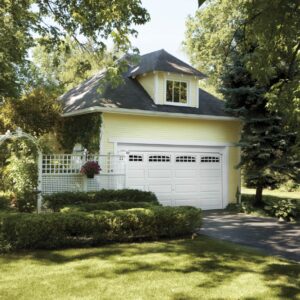 yellow home surrounded by a lot of shrubbery that has a clopay premium classic series, traditional style residential garage door