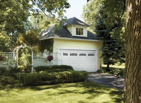 yellow home surrounded by a lot of shrubbery that has a clopay premium classic series, traditional style residential garage door