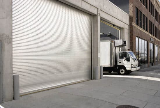two large commercial rolling garage doors, one closed and the other open with a white delivery type truck pulling out of it on the exterior