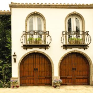 two arched double door custom wood residential garage doors positioned under two arched windows with balcony's that have plants on them