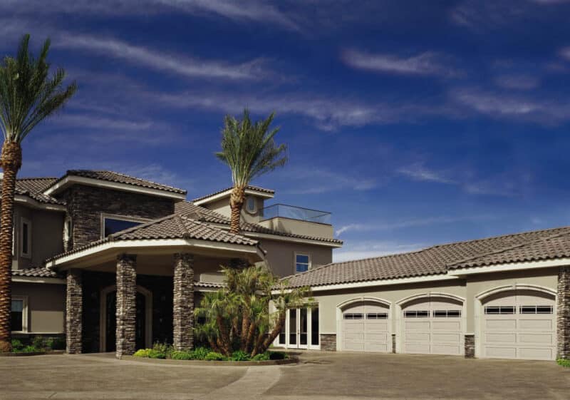 zoomed out view of a very large san antonio home that has three traditional style, colonial/ranch raised panel residential garage doors, palm trees around the house with a tiled roof