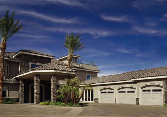 zoomed out view of a large san antonio home with grey roof tiles and three cream colored carriage style sonoma/sonoma ranch panel garage doors that have windows on the top row of the door