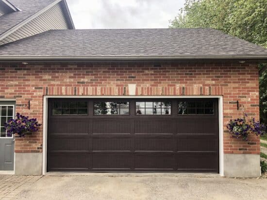 A neat residential garage with a dark brown door featuring glass windows at the top. The facade is made of red brick, and there are decorative flower pots hanging on either side, adding a touch of color and charm to the home's exterior