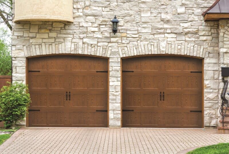 A luxurious home exterior with two large wooden garage doors set against a stone wall facade. The doors feature arched tops and traditional panel design, complemented by small black decorative handles and a classic wall-mounted lantern between them