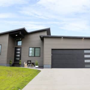 A modern single-story house with a dark gray facade and multiple angled rooflines, featuring large windows, a sleek black garage door with horizontal panels and glass inserts, and a welcoming porch area with a seating arrangement. The house is set against a backdrop of lush greenery and has a concrete driveway