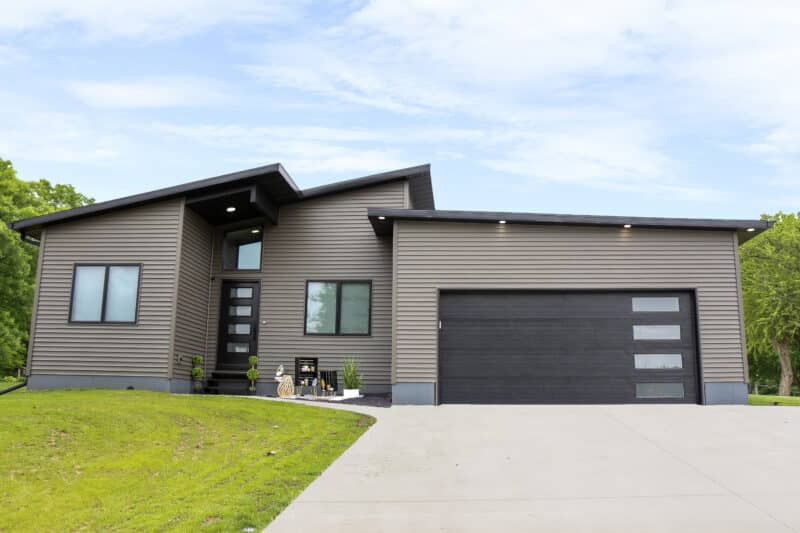 A modern single-story house with a dark gray facade and multiple angled rooflines, featuring large windows, a sleek black garage door with horizontal panels and glass inserts, and a welcoming porch area with a seating arrangement. The house is set against a backdrop of lush greenery and has a concrete driveway