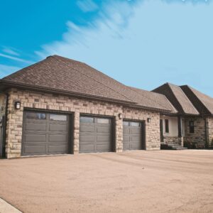 A spacious stone-covered house featuring a hip roof with asphalt shingles and three gray garage doors that blend seamlessly with the facade. The home includes multiple windows, and a gravel driveway leads up to the garages, situated in a well-manicured suburban setting under a clear blue sky