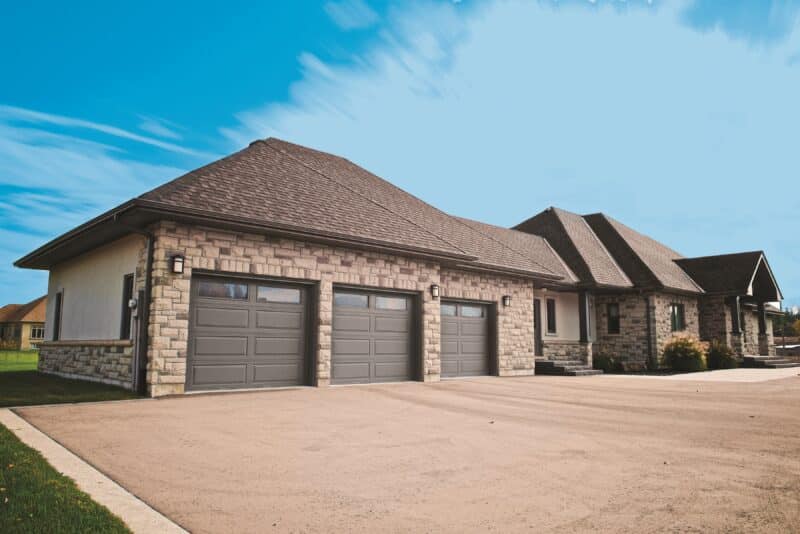 A spacious stone-covered house featuring a hip roof with asphalt shingles and three gray garage doors that blend seamlessly with the facade. The home includes multiple windows, and a gravel driveway leads up to the garages, situated in a well-manicured suburban setting under a clear blue sky