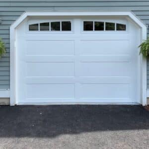 A clean, modern single white garage door with a rectangular design, featuring small windows along the top. The garage door is set in a light blue wooden siding of a house, flanked by black lantern-style sconces and hanging green plants on both sides