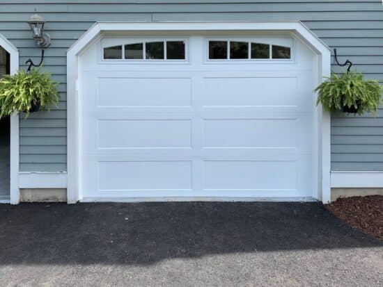 A clean, modern single white garage door with a rectangular design, featuring small windows along the top. The garage door is set in a light blue wooden siding of a house, flanked by black lantern-style sconces and hanging green plants on both sides