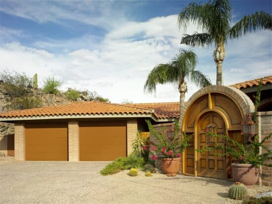 Southwestern style home with terracotta roof and three tan garage doors, complemented by a desert landscape with cacti and palm trees