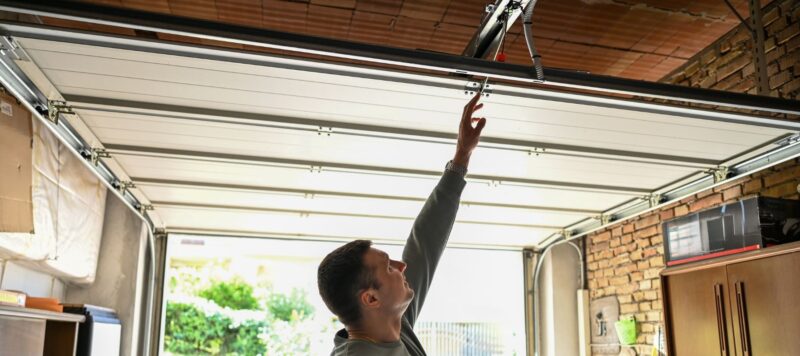 A man reaches up to inspect a garage door opener track and the top panel of an open garage door inside a garage with brick walls